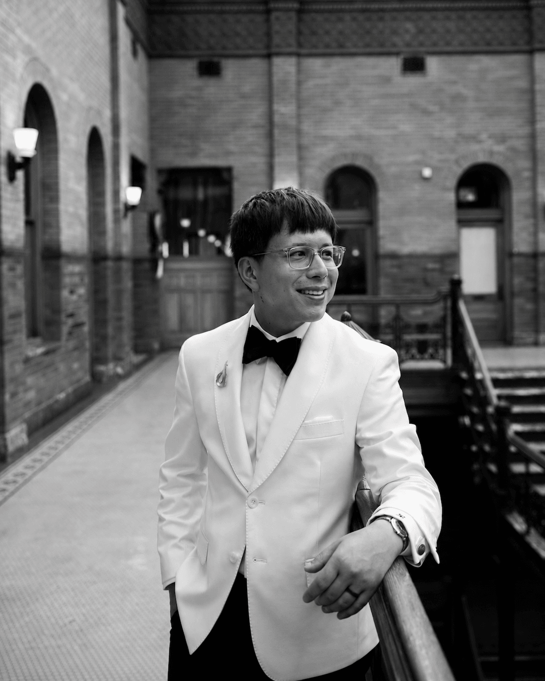 Person in a tuxedo with a white jacket and black bow tie, smiling, standing outdoors near a railing.
