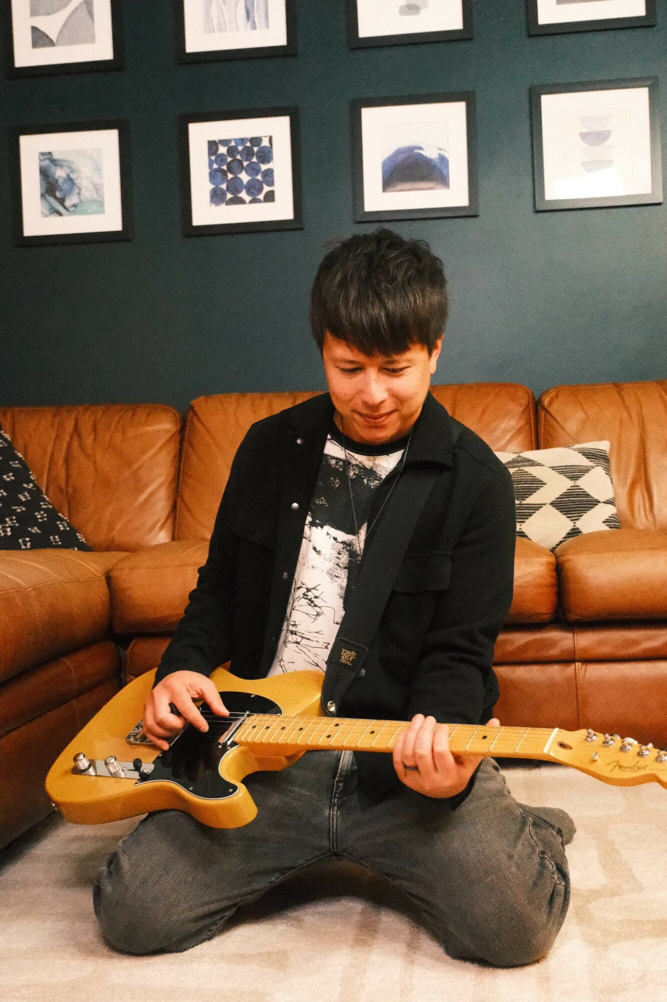 Young person sitting on floor playing yellow electric guitar, brown leather sofa and framed pictures on dark green wall behind.