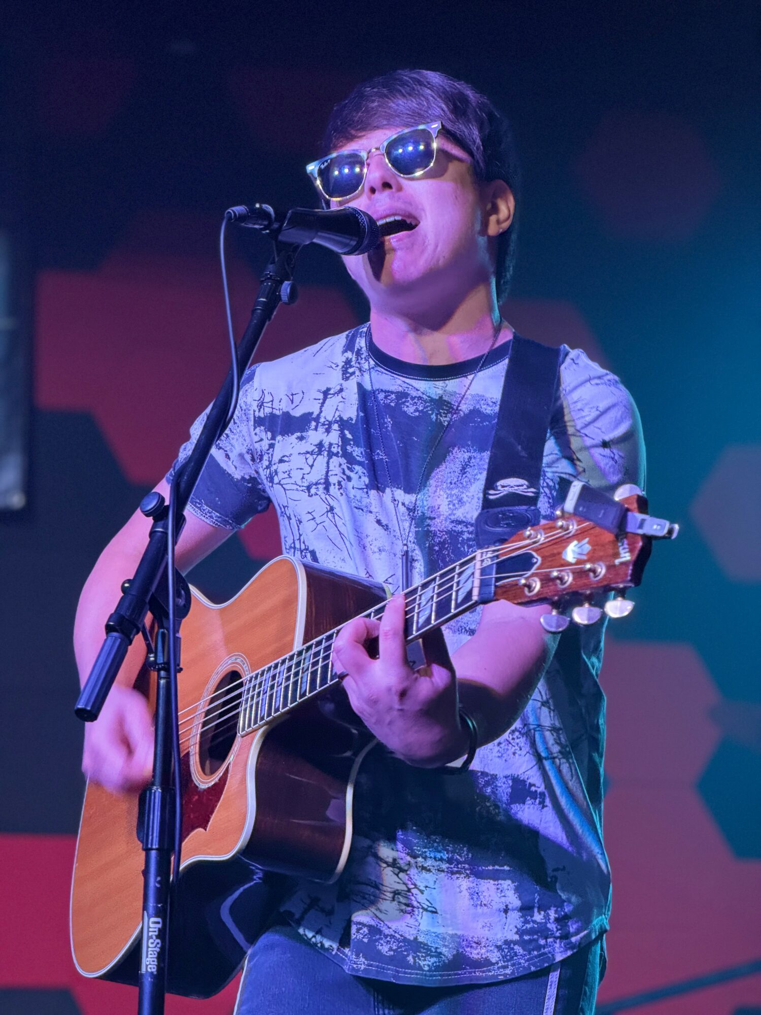 Young person wearing sunglasses, singing into a microphone, playing an acoustic guitar on stage with colorful lighting.
