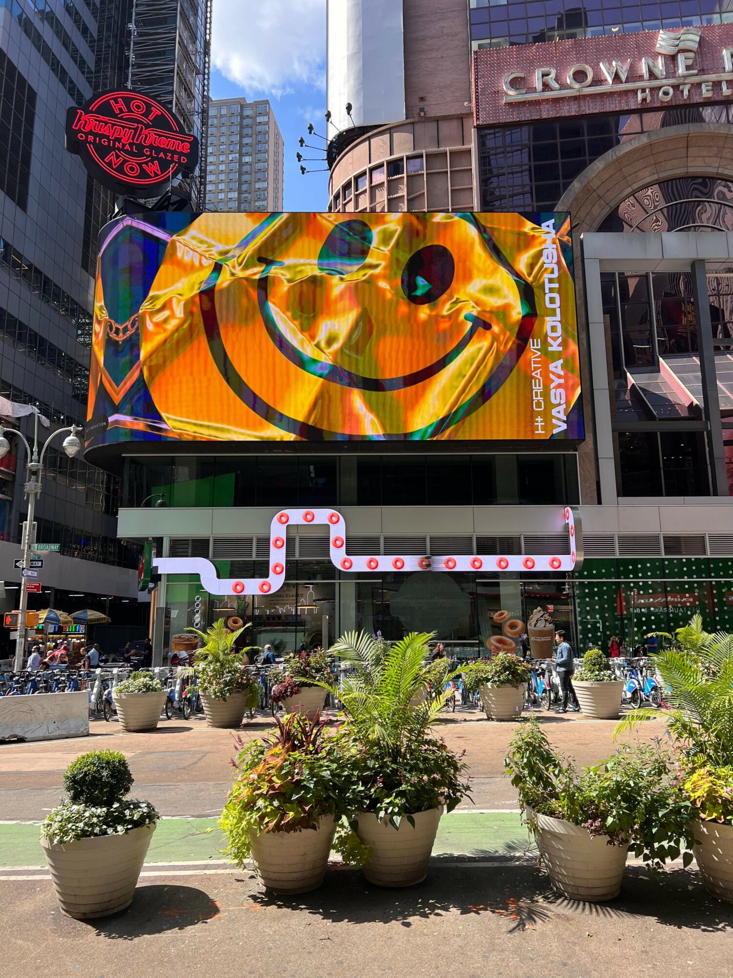 City street scene with a large colorful advertisement, potted plants, and people walking on the sidewalk.