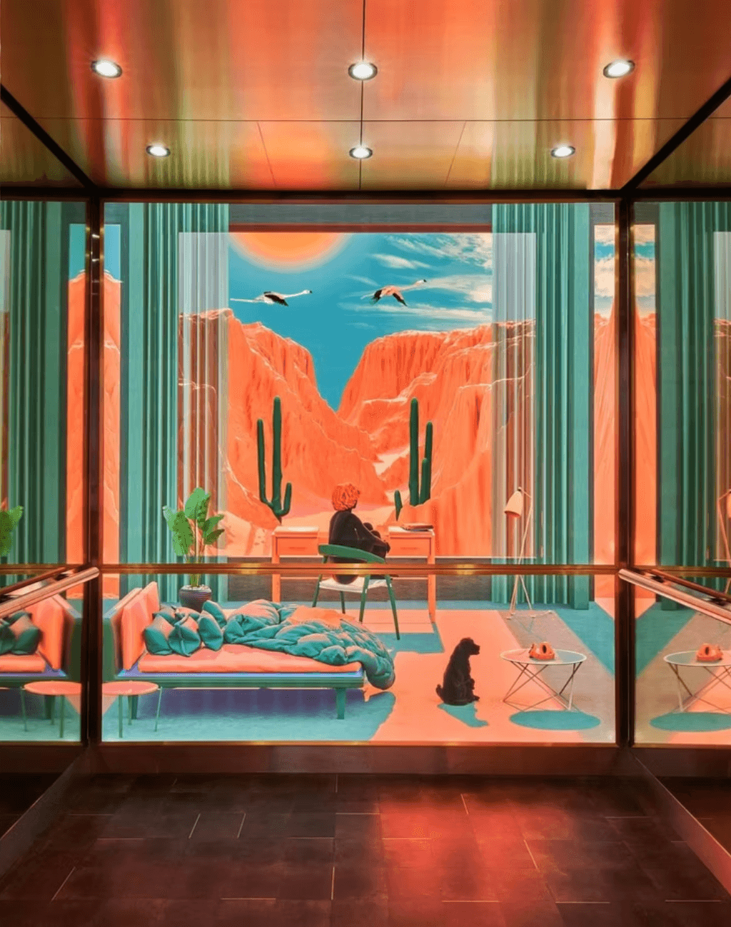 Room with desert mural, person sitting at desk, sofa with blanket, cat, and small table, viewed through glass with railing.