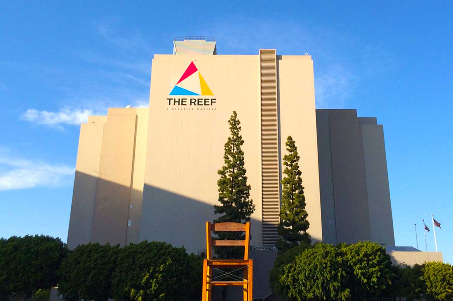 Large building with a sign reading 'THE REEF' and a colorful logo, two tall trees in front, and bushes at the base.