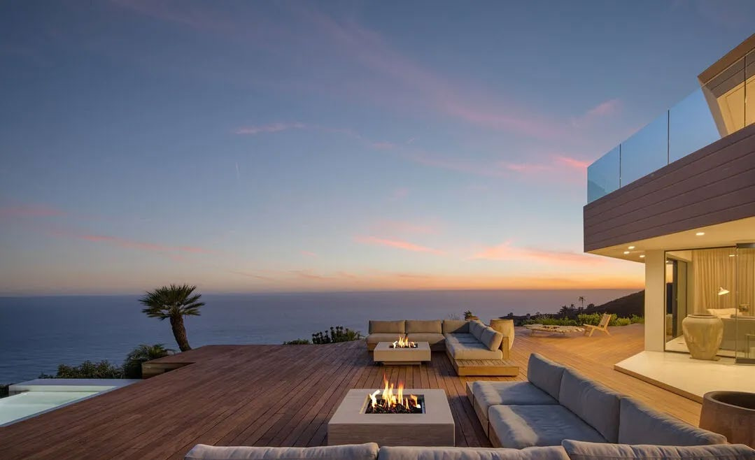 Outdoor patio with seating area overlooking ocean and sunset sky, adjacent to modern house with glass balcony.