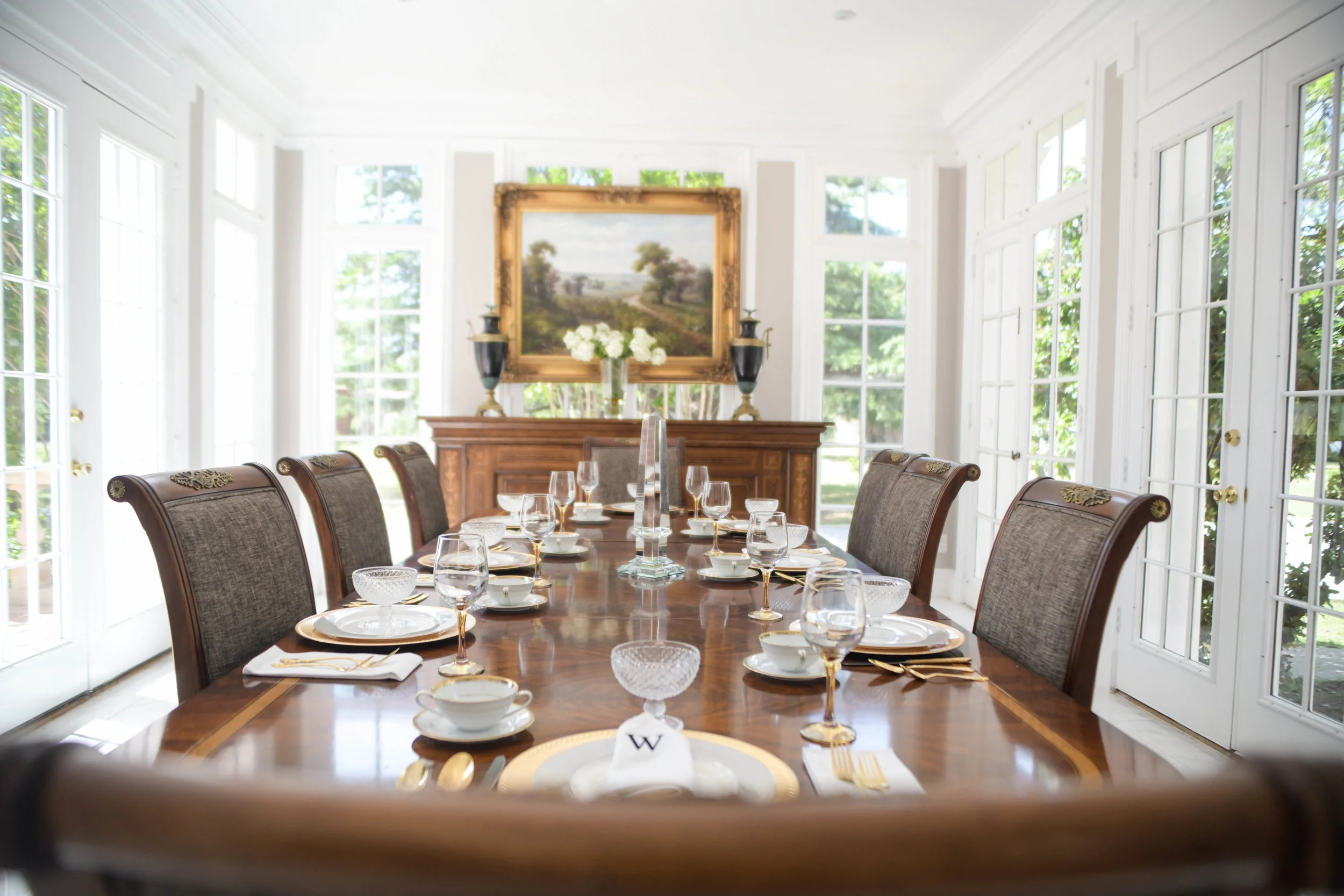 Elegant dining room with a long wooden table set with plates, glasses, and napkins, surrounded by upholstered chairs, bright windows, and a painting.