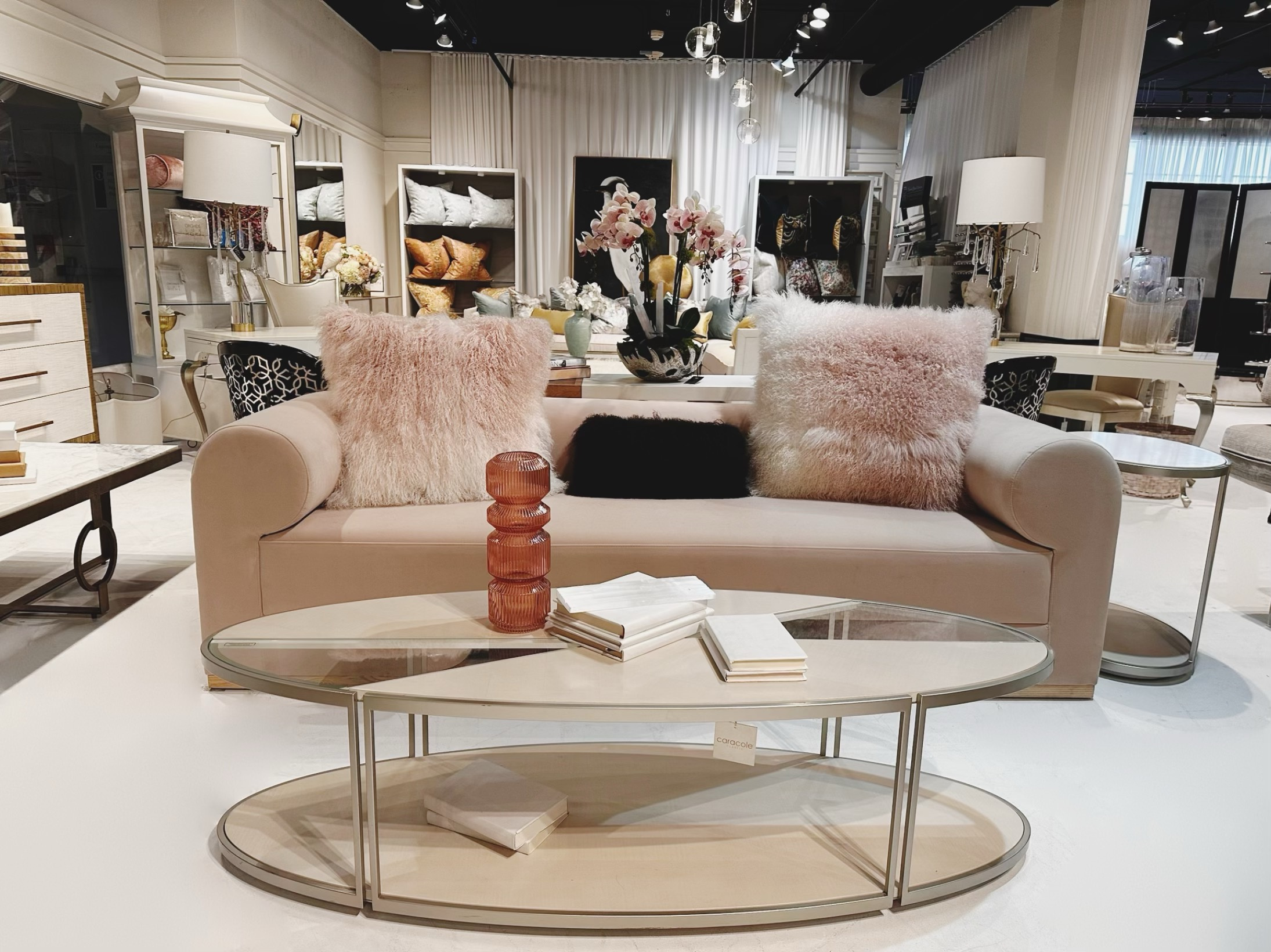 A beige sofa with two fluffy pink pillows, a glass coffee table with stacked objects, in a well-lit furniture store.