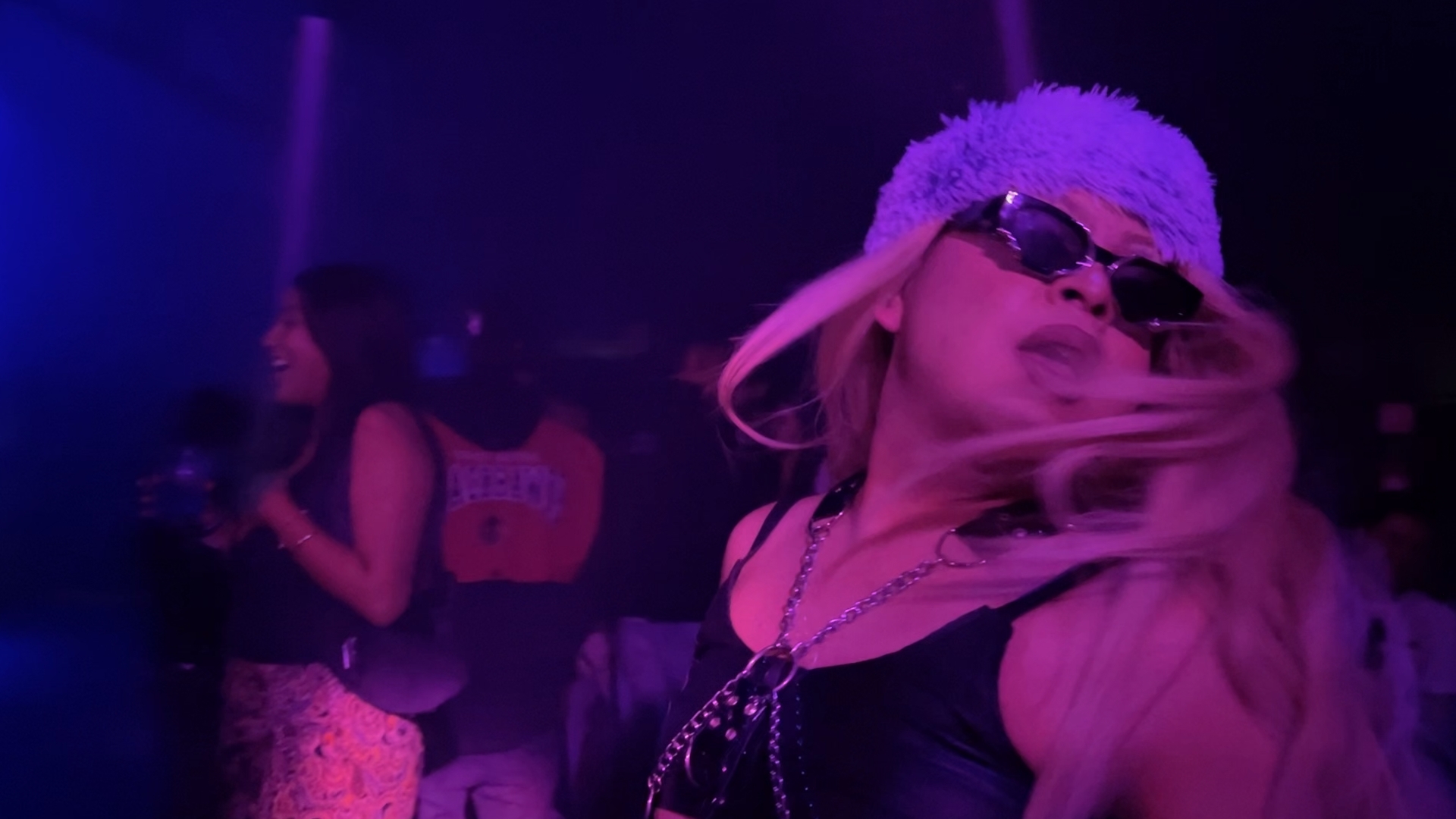 Person wearing sunglasses and a hat with long hair, in a dark, colorful party setting.