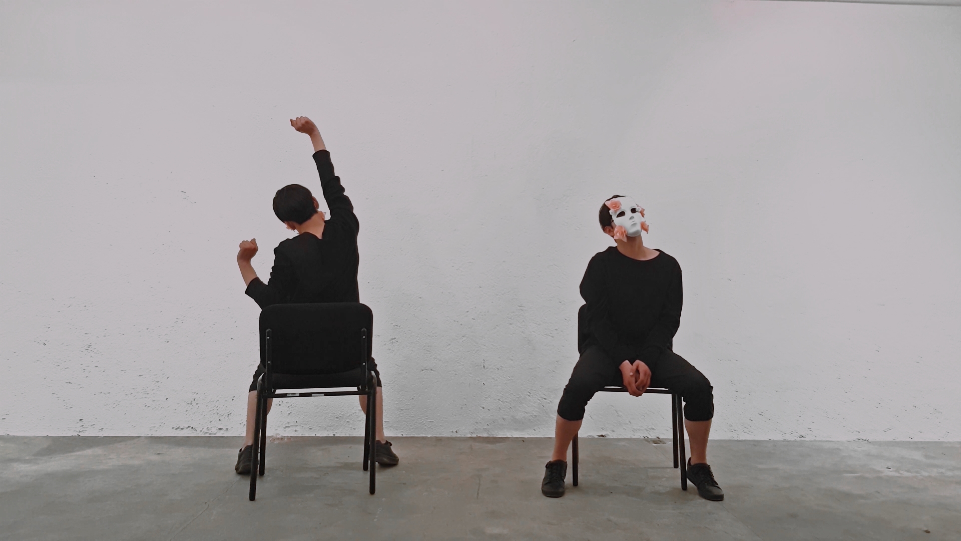 Two people sit on chairs against a plain wall, one with a mask, the other with their back to the camera.