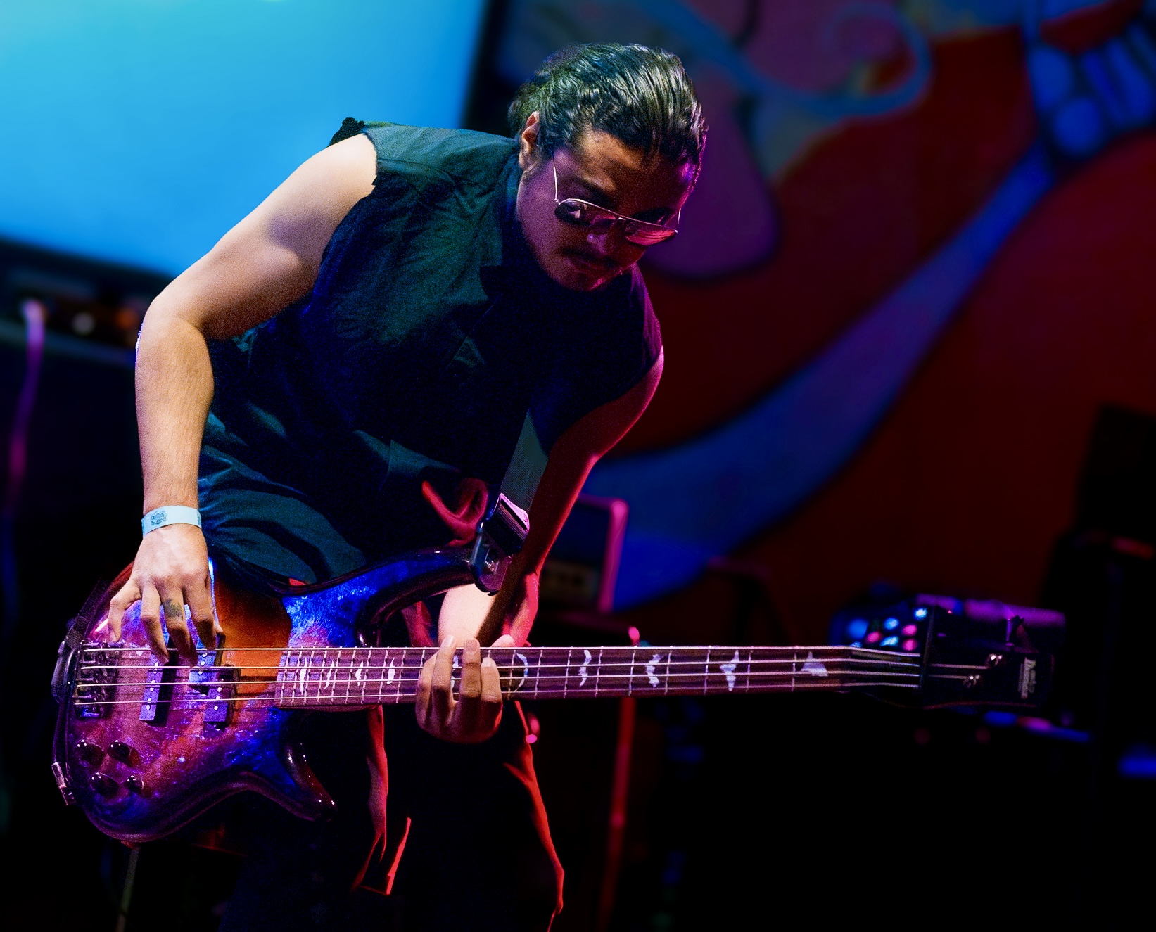 Person playing an electric guitar on stage, wearing glasses and a sleeveless top, with colorful background lighting.