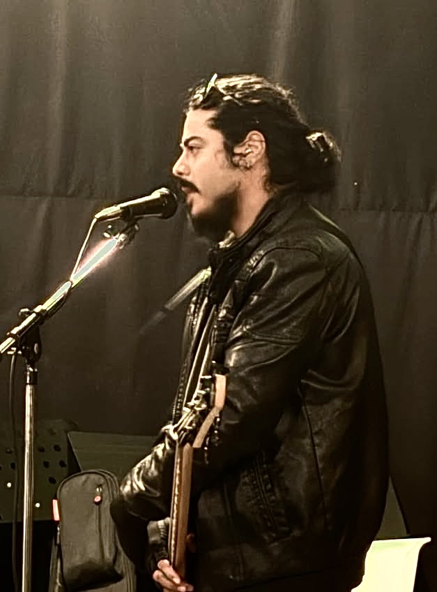 Person with long dark hair and beard singing into a microphone, wearing a black leather jacket, holding a guitar, on stage.