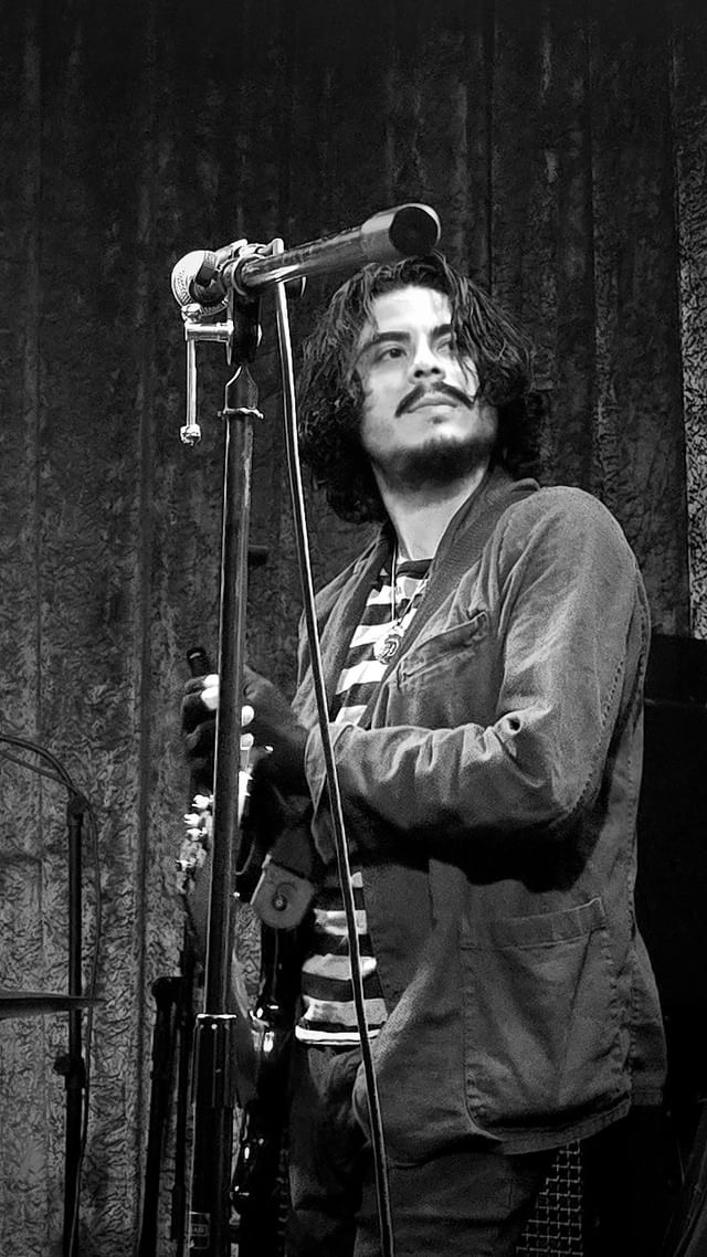 Person with long hair and beard standing on stage, holding a microphone, in front of a dark curtain.