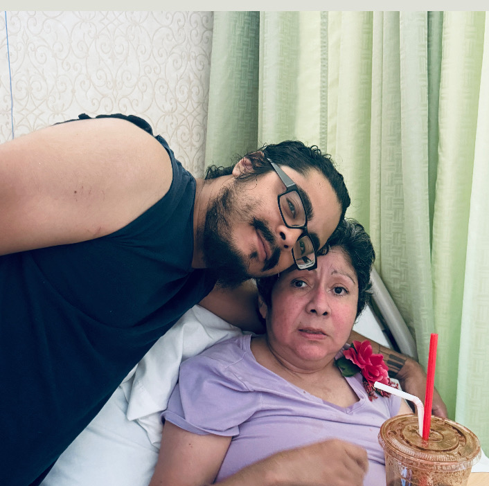 A man with glasses leans over a woman in a hospital bed with a cake and candle, green curtains in background.