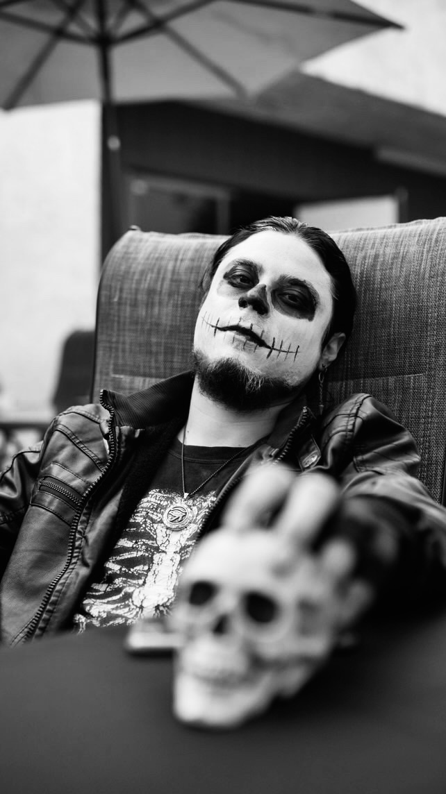 Person with face paint resembling a skeleton, sitting at a table with a skull prop, outdoors, black and white photo.