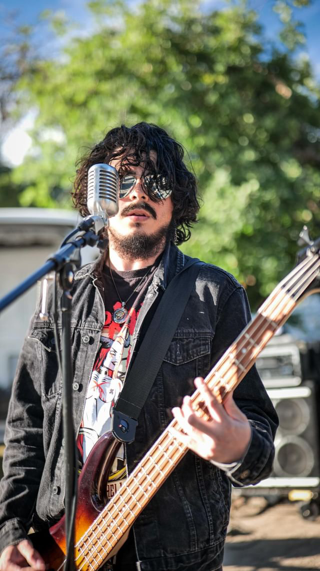 Person with curly hair and sunglasses playing a bass guitar outdoors, with trees and a microphone nearby.