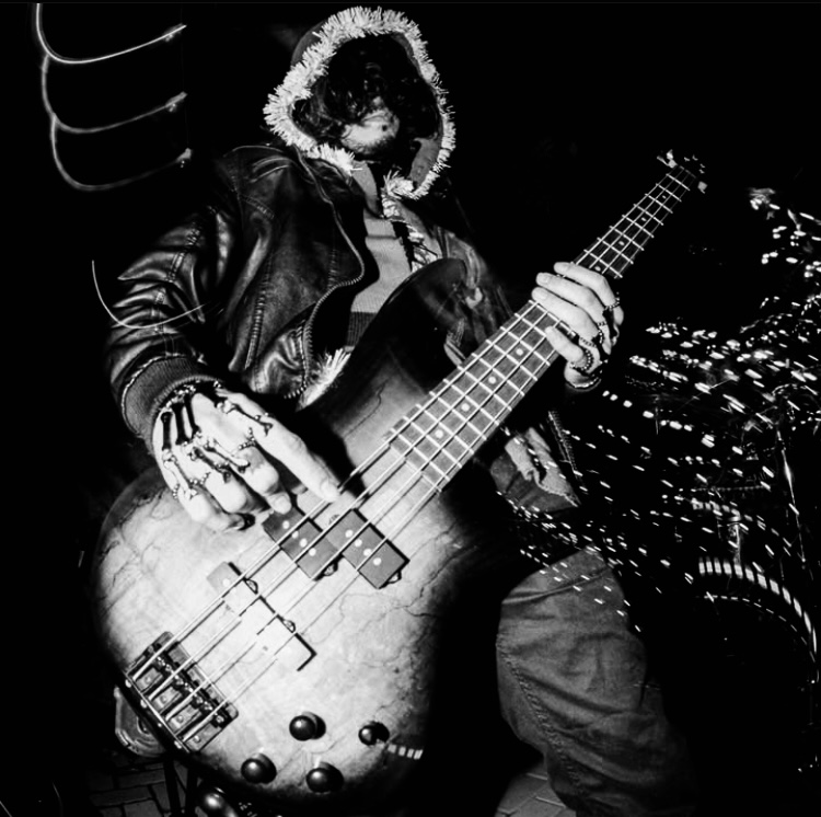 Person wearing a hoodie and leather jacket playing a bass guitar at night, city lights in background, black and white.