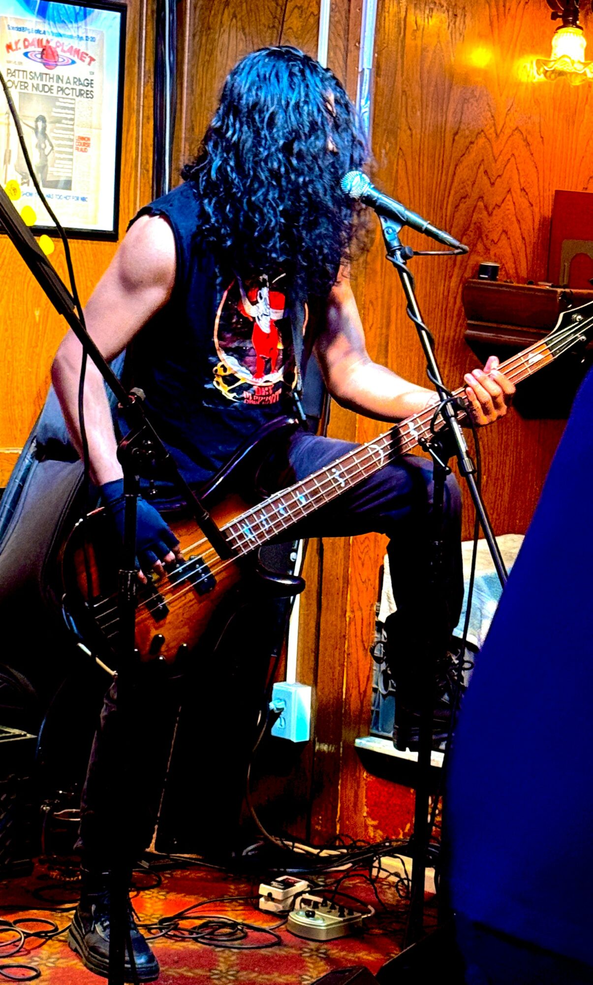 Person with long curly hair playing bass guitar and singing into a microphone in a wooden-paneled room.