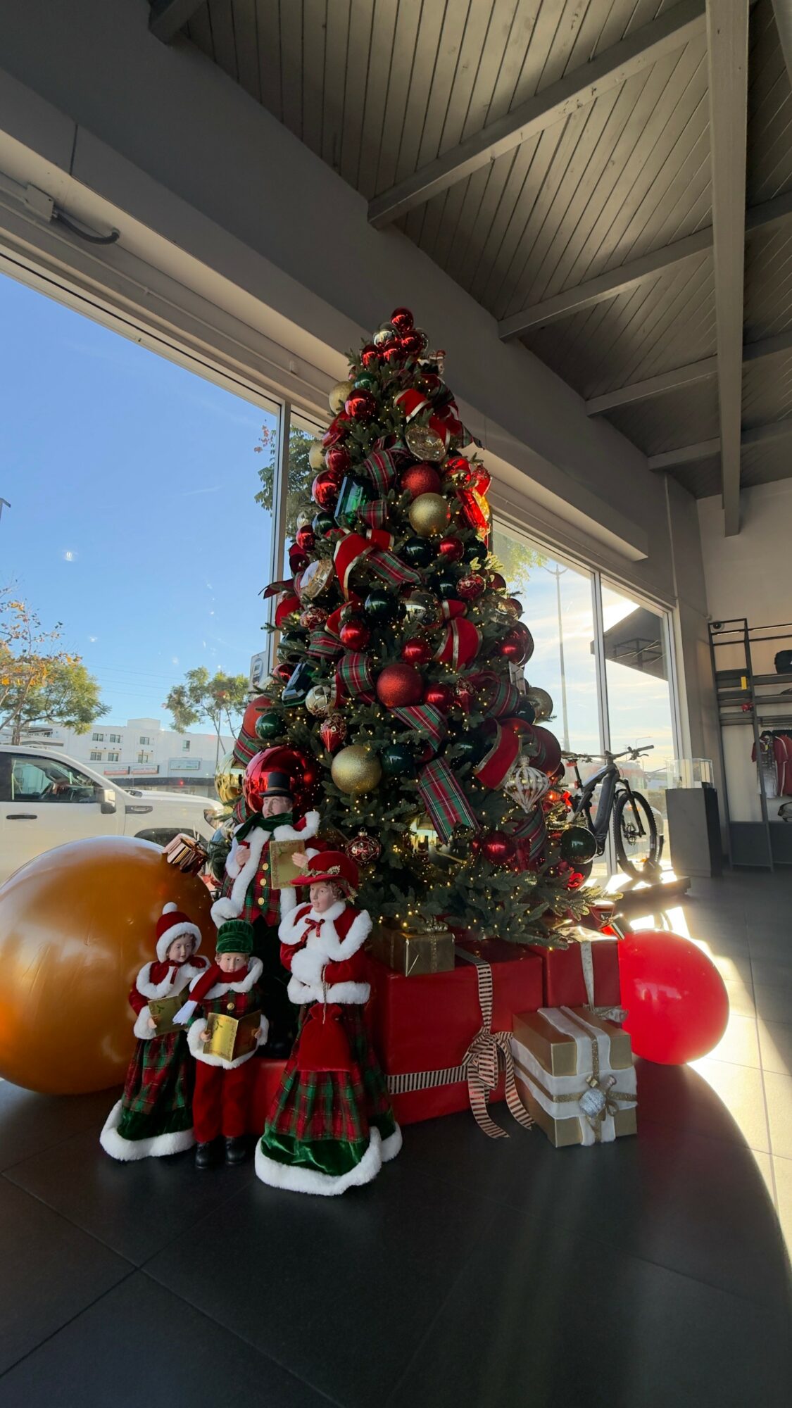 Decorated Christmas tree with presents and festive figures indoors, with large window showing blue sky and trees outside.