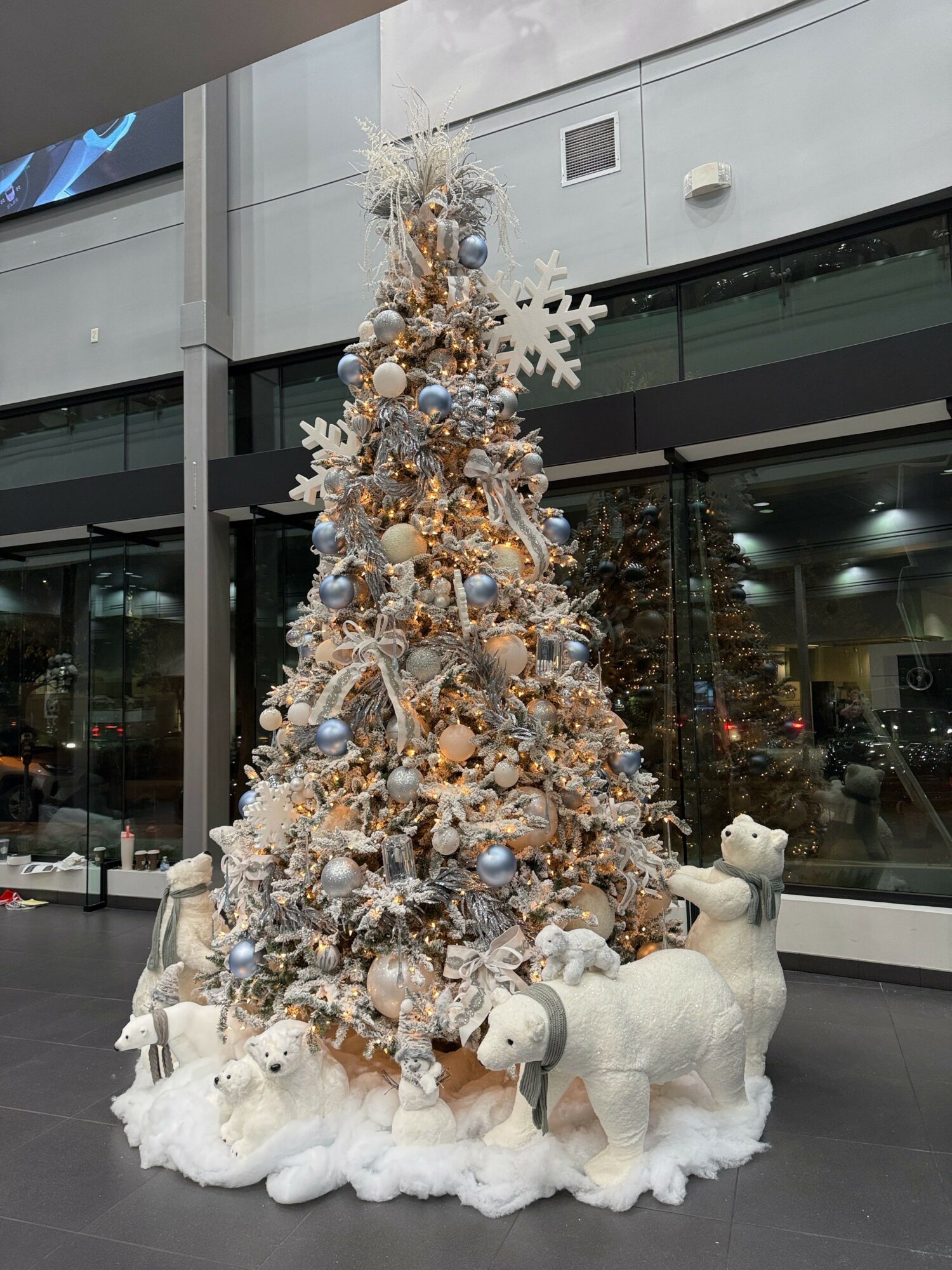 Decorated Christmas tree with ornaments, surrounded by white plush animals, in front of a glass window.