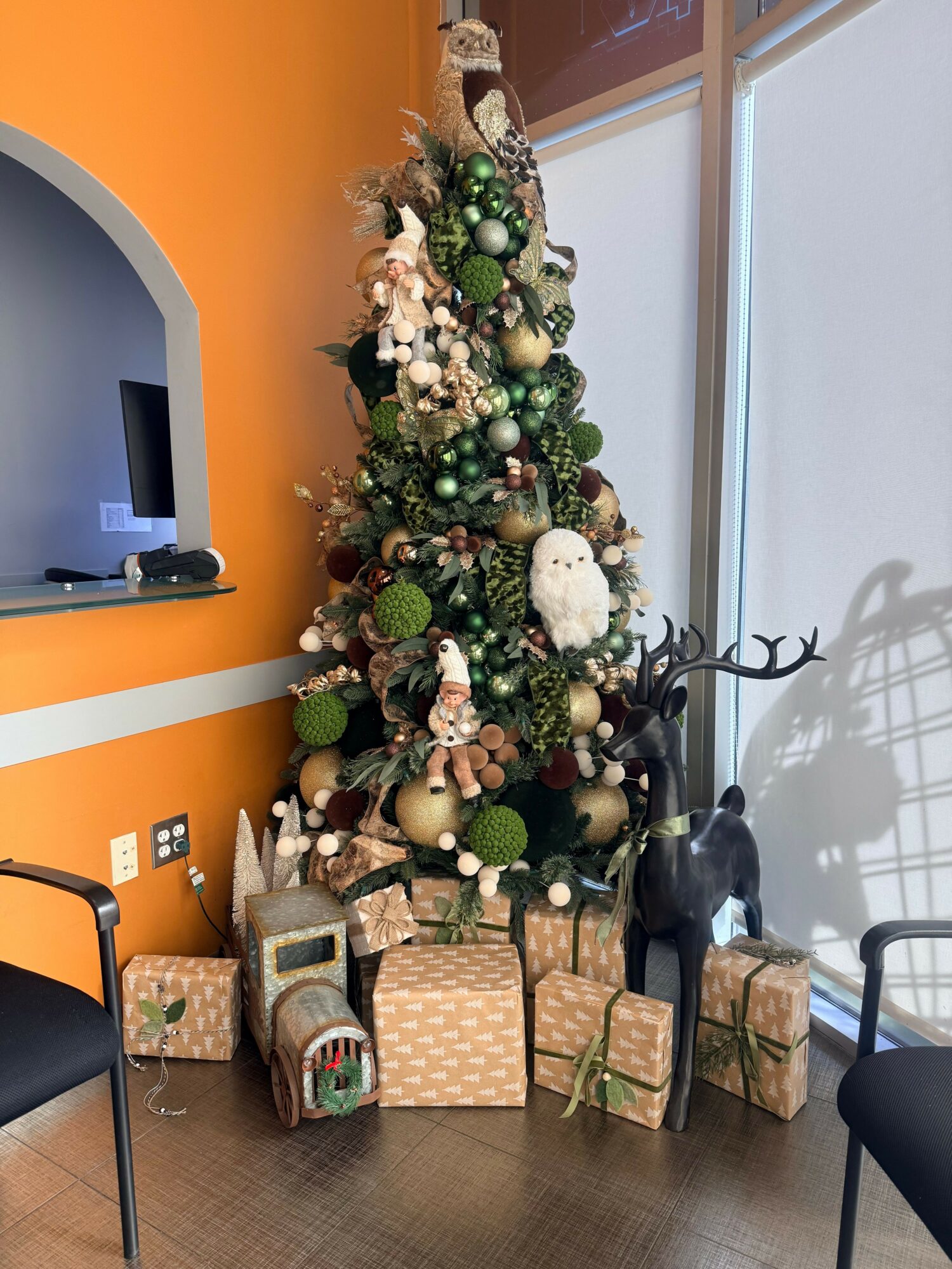 Decorated Christmas tree with ornaments, gifts, and a reindeer figure, placed indoors against an orange wall.