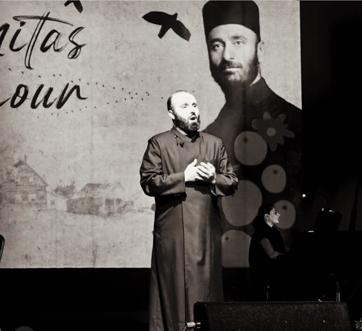 Man in traditional attire standing on stage with a large portrait of a man in a hat projected behind him, and birds flying in the background.