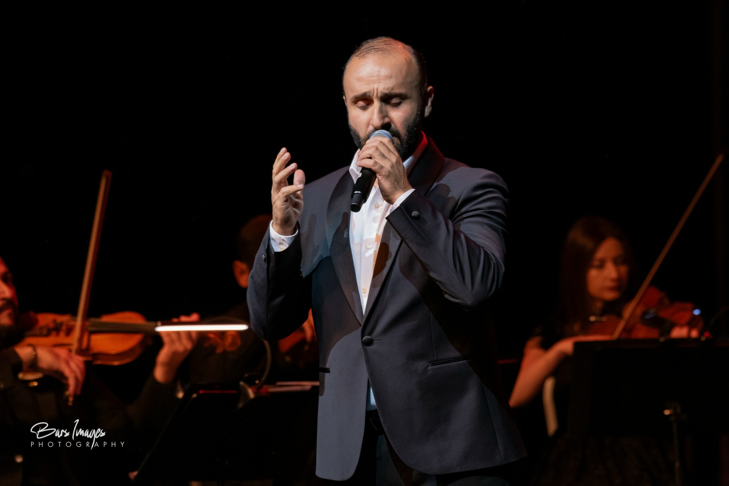 Man in suit singing into microphone with orchestra in background, stage lighting, black background