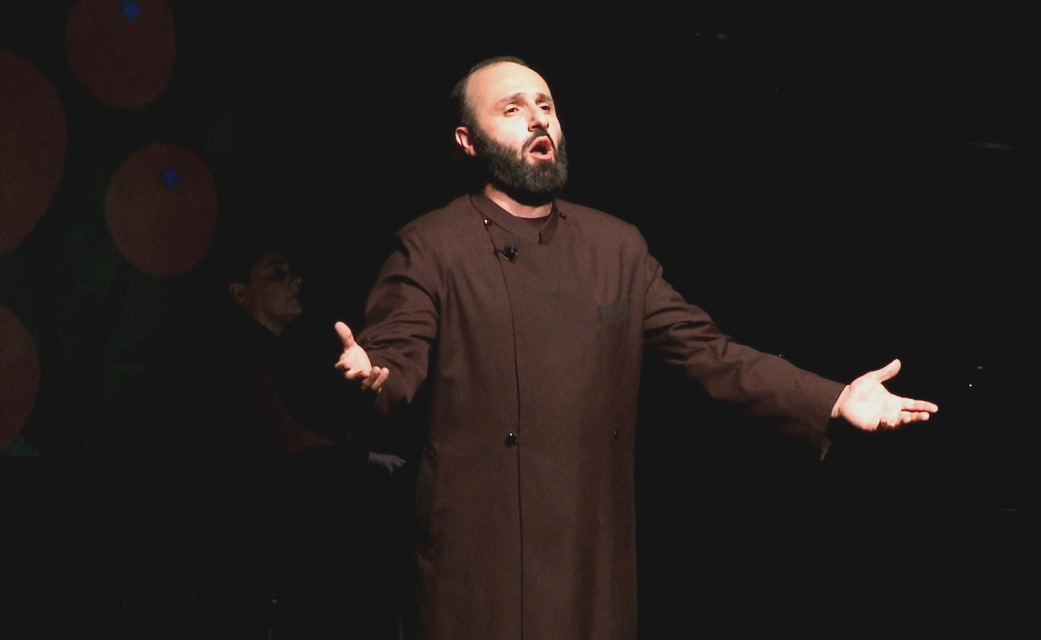 Man with beard and dark clothing performs on stage with arms outstretched, dark background, stage lighting.