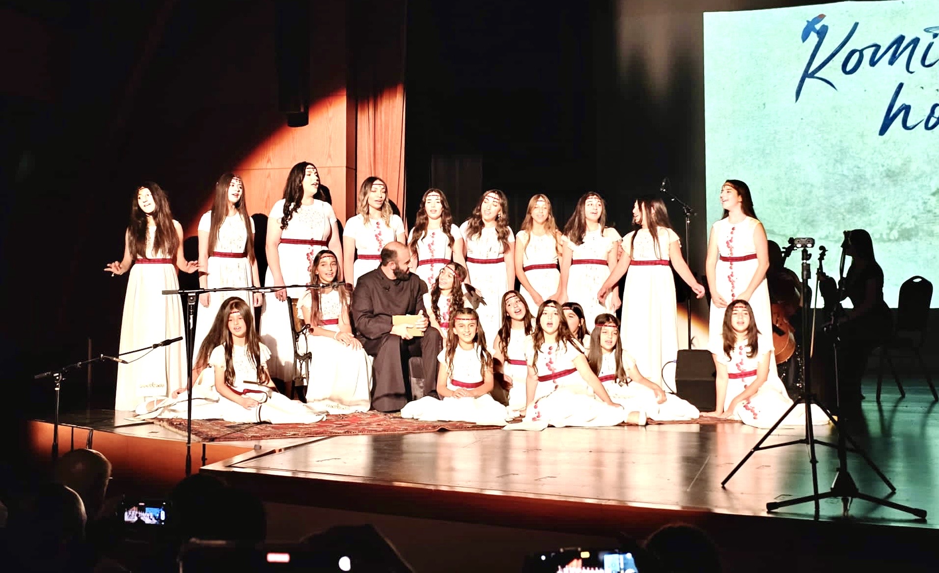 Group of young girls in white dresses with red sashes on stage, with a conductor in front, and a screen in the background.