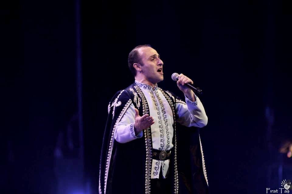 A man singing into a microphone on stage, wearing traditional clothing, with a dark background.