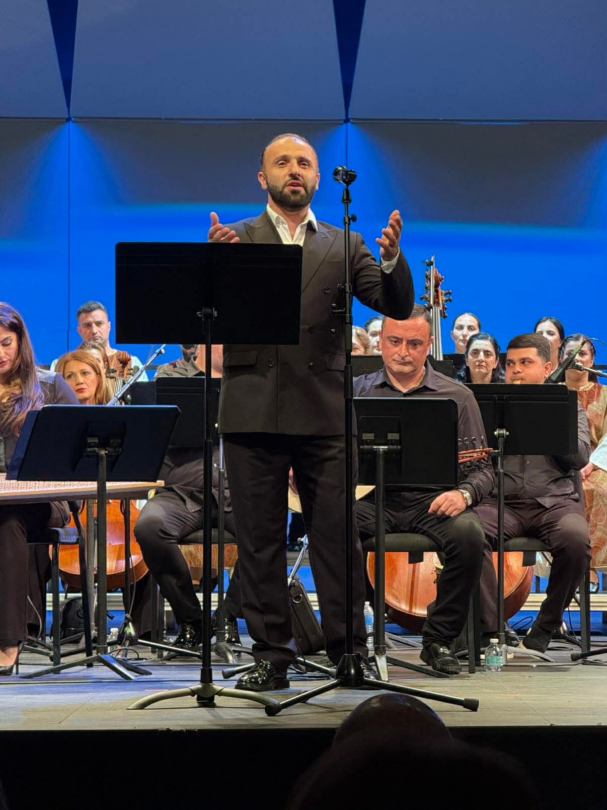 Conductor stands in front of orchestra on stage, gesturing with hands, with musicians and music stands behind him.