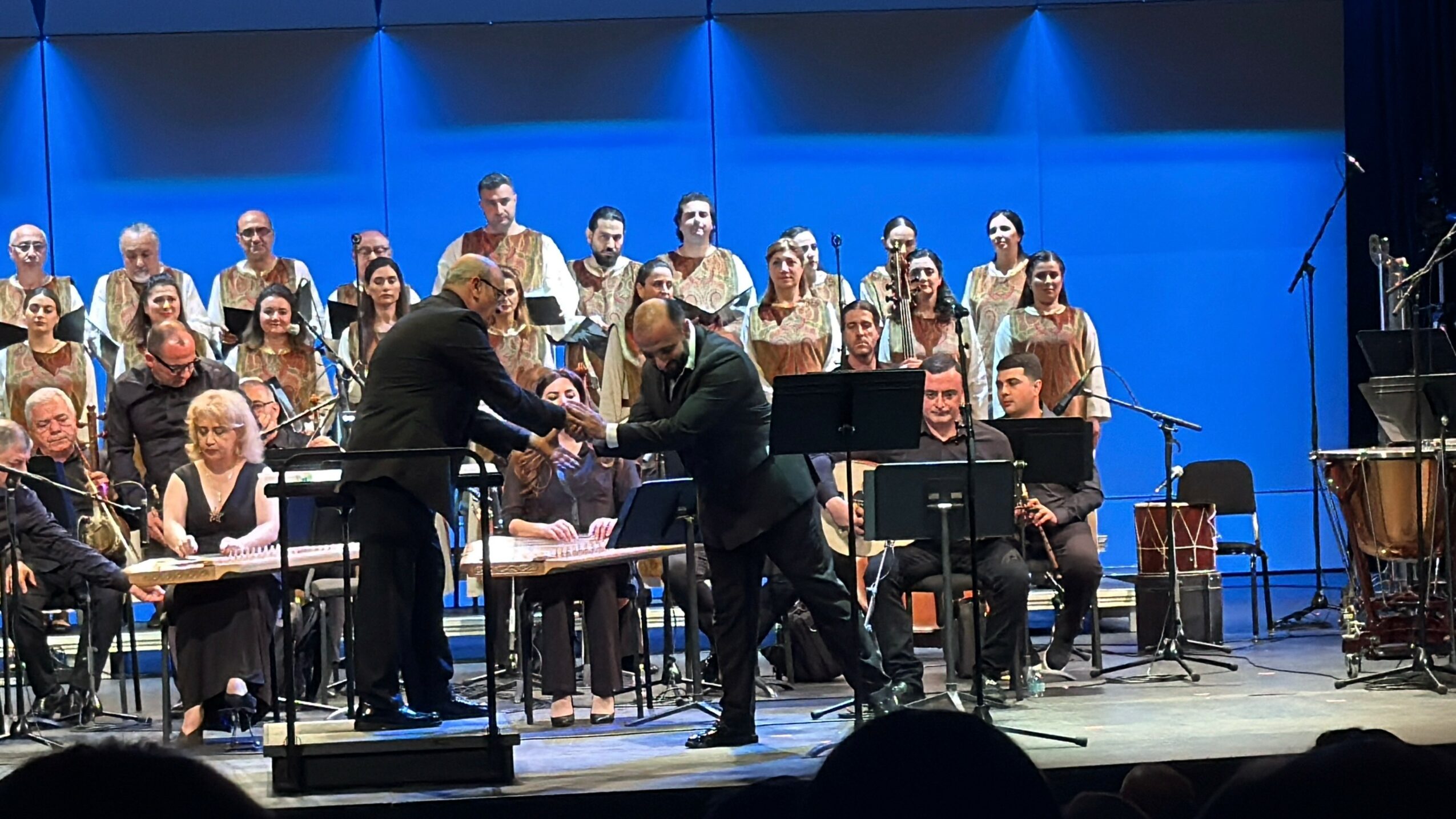 Orchestra and choir performing on stage with two people exchanging a document, blue background, musicians seated with instruments.