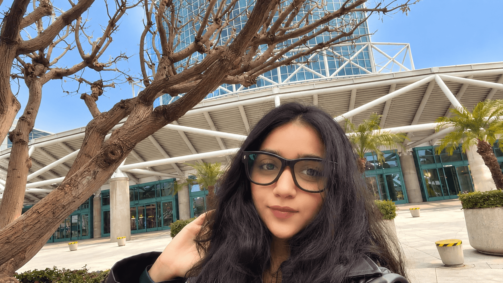 Young woman with glasses and long dark hair outdoors near a modern building and tree.