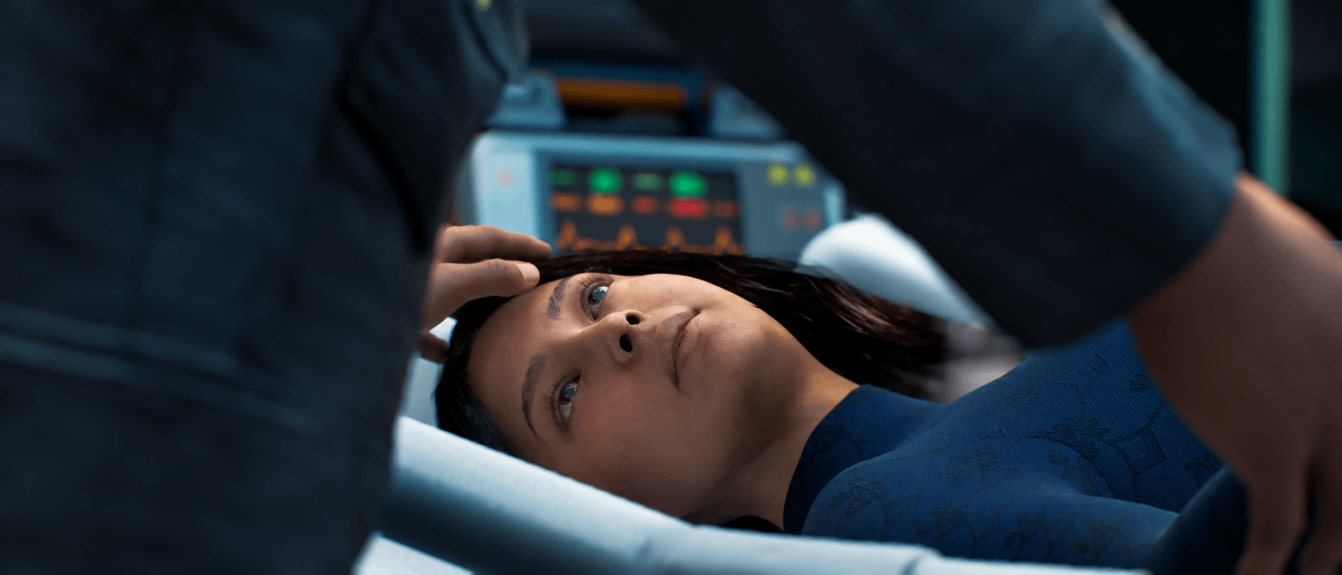 Person lying in a medical setting, looking up at a healthcare professional, with medical equipment in the background.