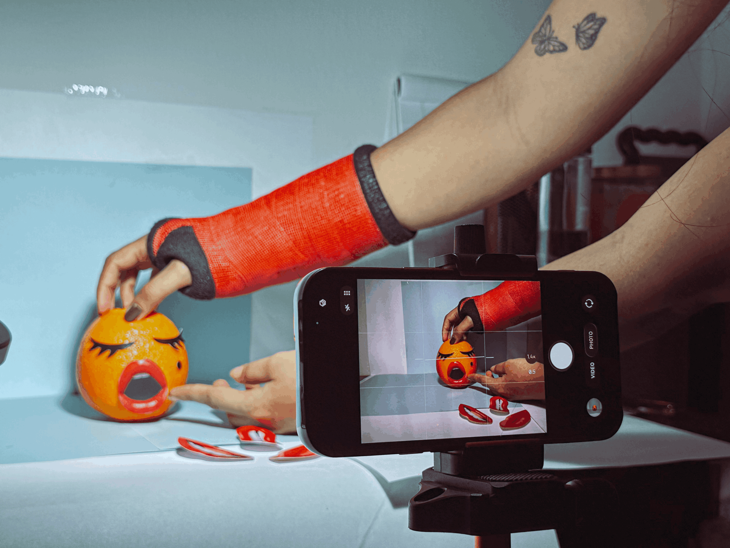 Person's arm with a red sleeve reaching for a yellow emoji-shaped container on a table, with a smartphone recording the scene.