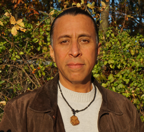 Man with dark hair and a serious expression stands outdoors with green foliage behind him.