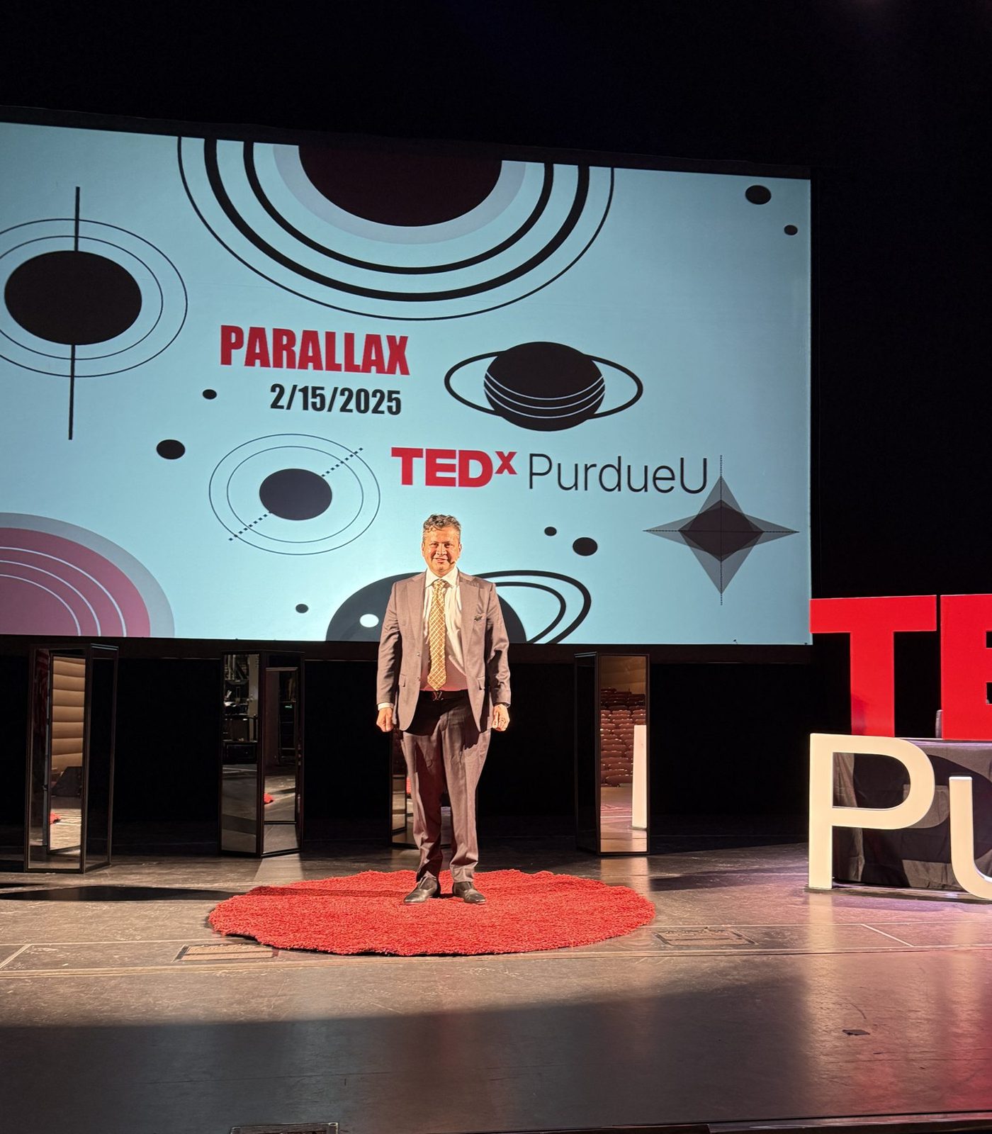 Man in suit standing on stage with TEDx Purdue logo and event details on screen behind him.
