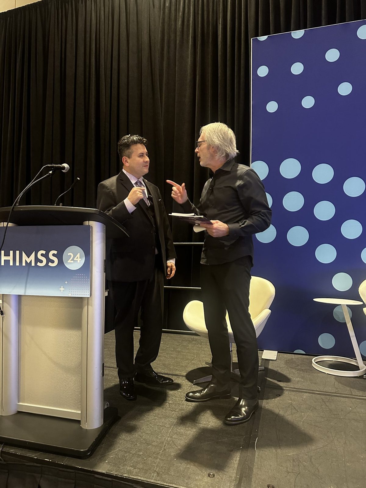 Two men are engaged in conversation on stage, with a podium and a large blue dotted background behind them.