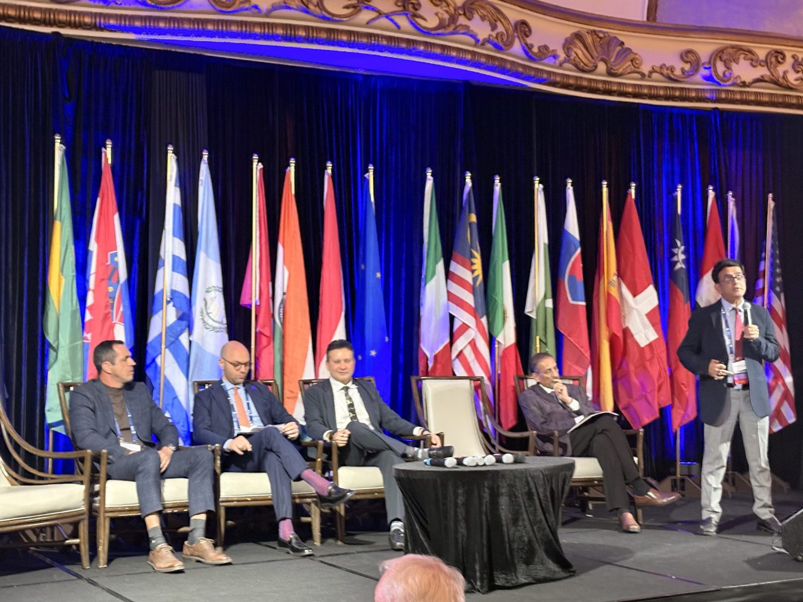 Five men seated on stage with a man standing and speaking, behind a row of international flags, in a formal setting.