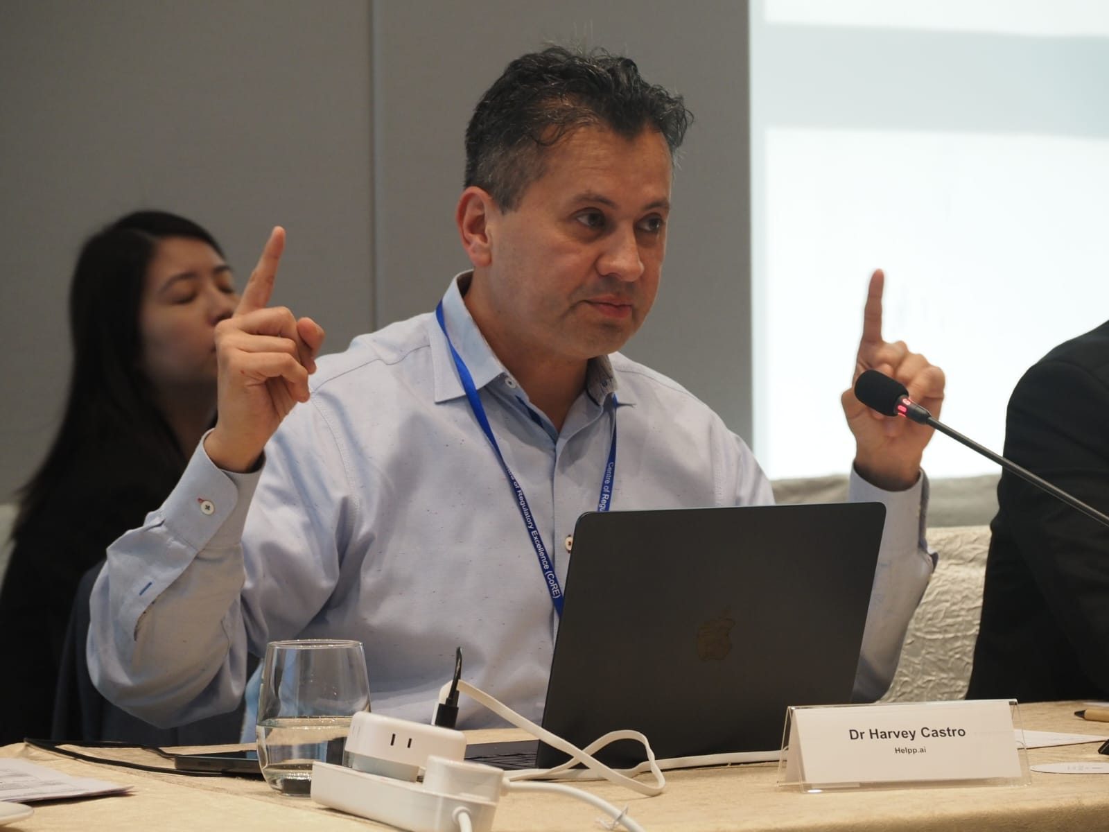 Man in white shirt with a laptop, raising both hands with index fingers up, in a meeting room.