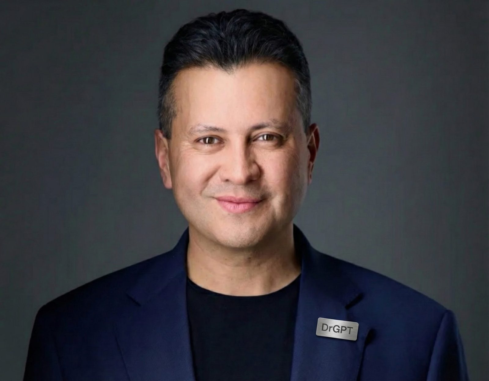 Portrait of a man with dark hair, wearing a dark blazer and a name tag, smiling against a dark background.