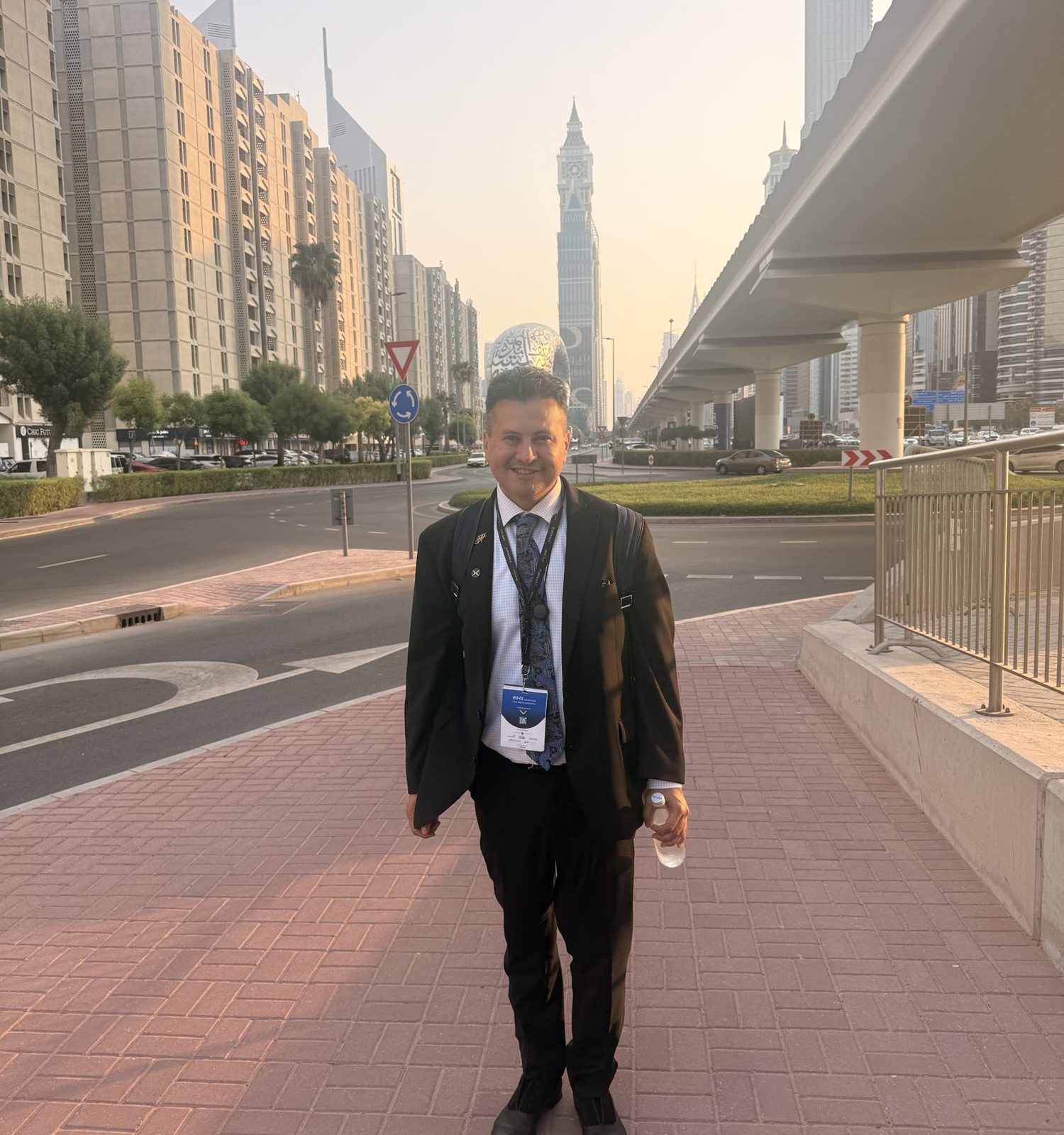 Man in suit standing on sidewalk with city buildings and a tall tower in background, holding a water bottle.