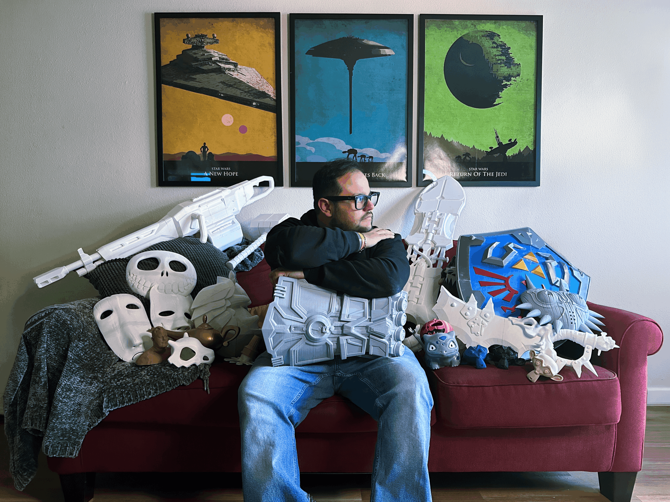 Man sitting on a red couch surrounded by masks and sci-fi props, with three framed space-themed posters on the wall behind.