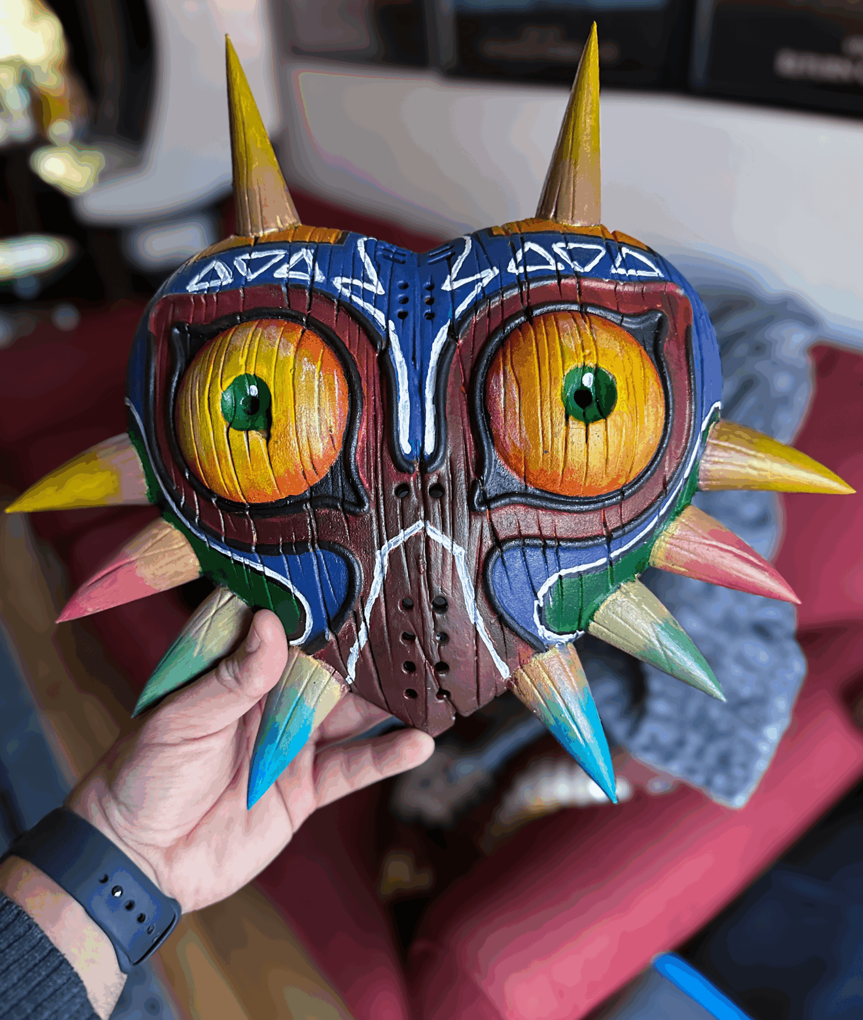 Hand holding a colorful, spiked mask resembling a face with large eyes and intricate patterns, indoors.
