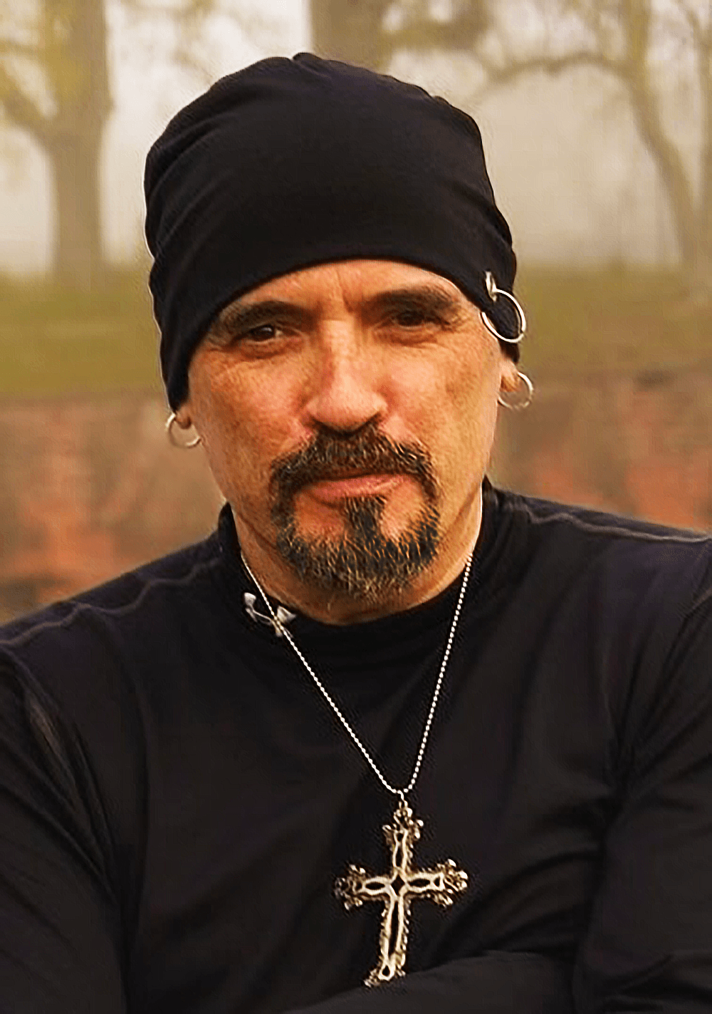 Man with a black head covering, earrings, and a cross necklace outdoors, looking at the camera.
