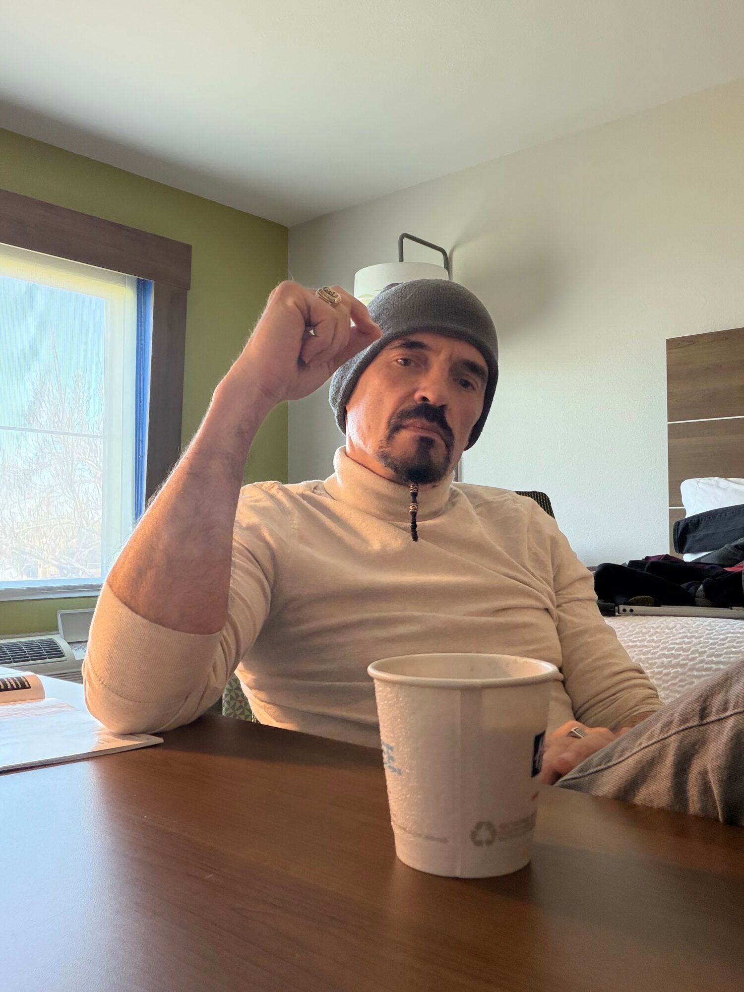 Man wearing a gray beanie and beige shirt sitting at a table with a paper cup, in a room with a window and a bed.