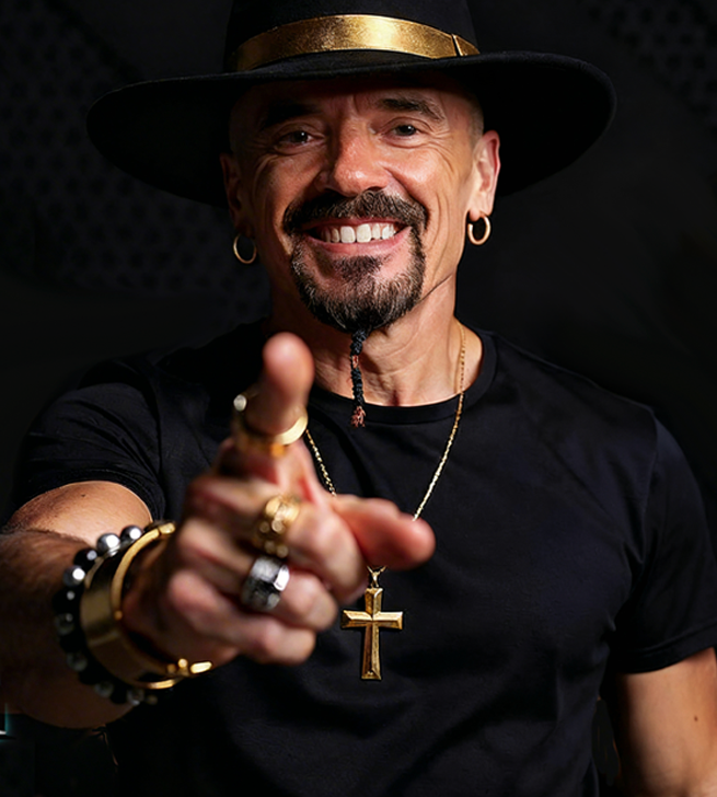 Smiling man wearing a wide-brimmed hat, gold jewelry, and a cross necklace, pointing towards the camera.