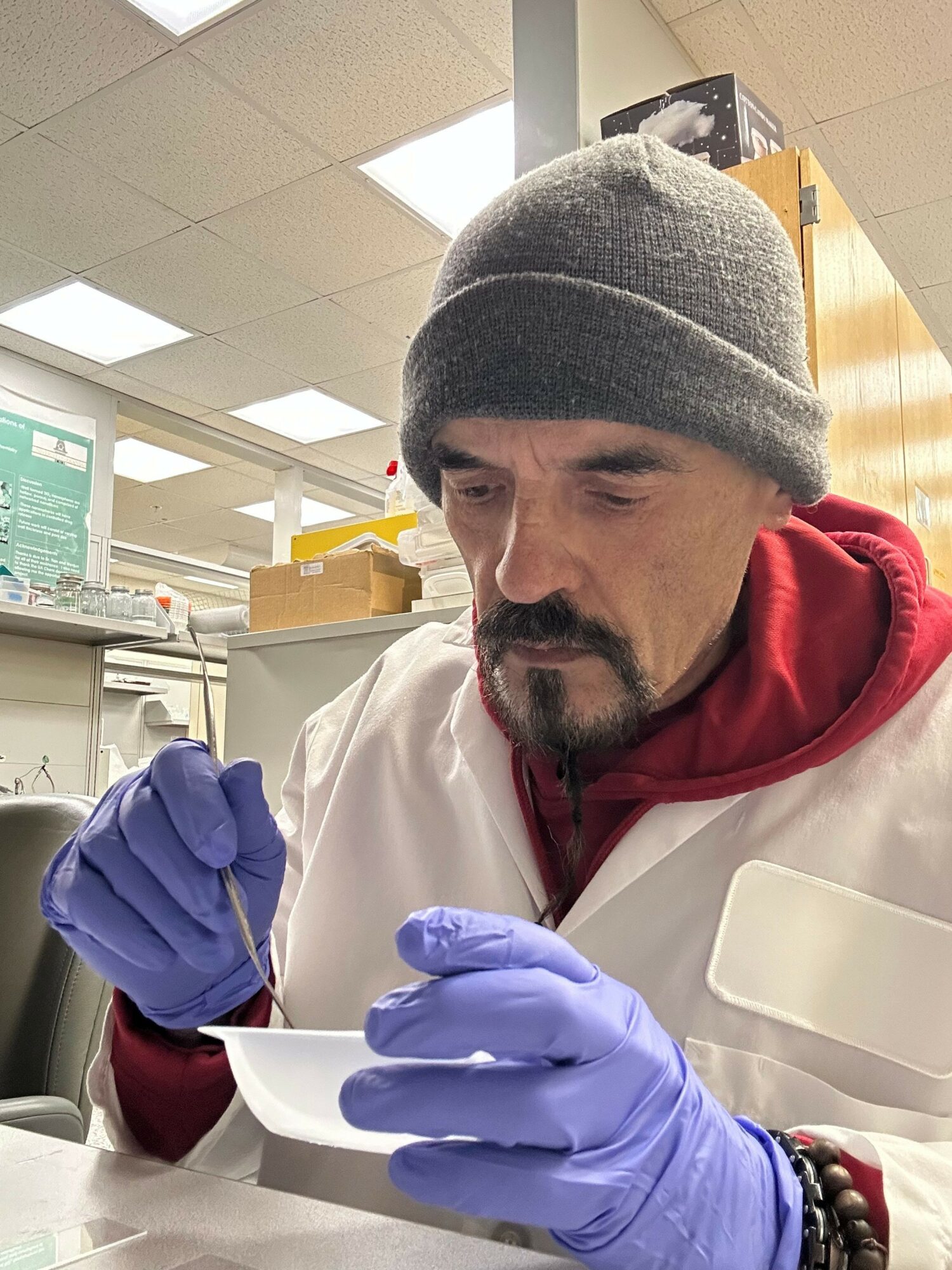 Man in a gray beanie and purple gloves working with a small tool at a table in a laboratory.