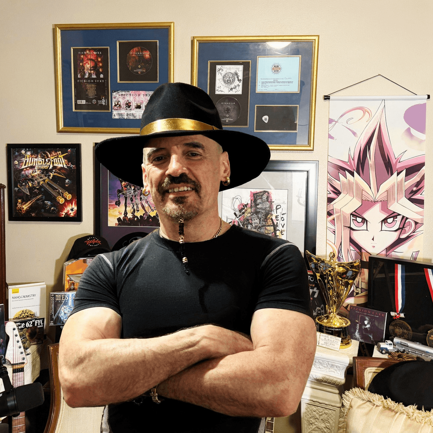 Man with a beard and mustache wearing a black hat with a gold band, standing with arms crossed in front of framed artwork and posters on a wall.