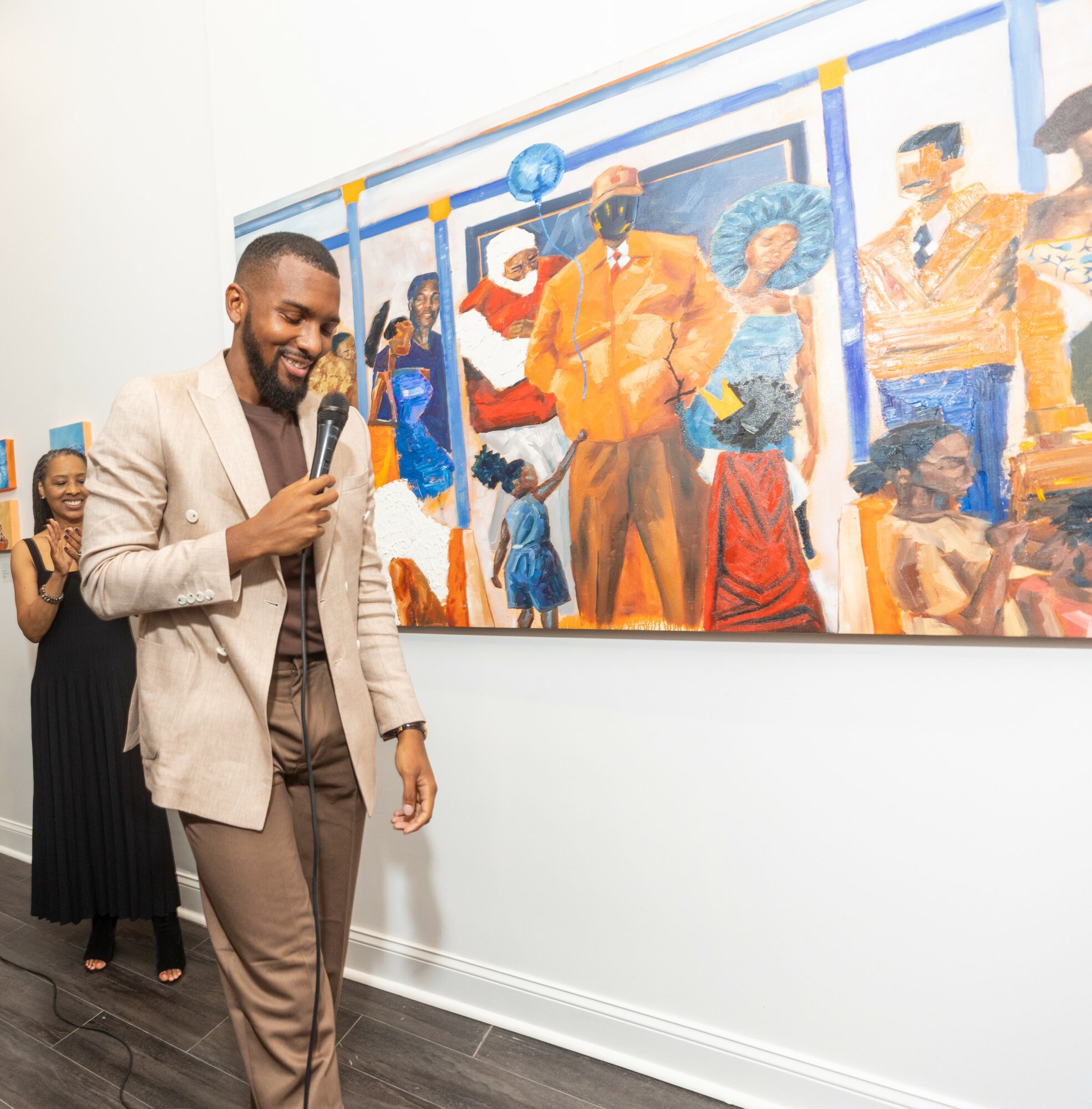 Man smiling and holding a microphone in front of a colorful mural, with two women in the background. Word count: 20