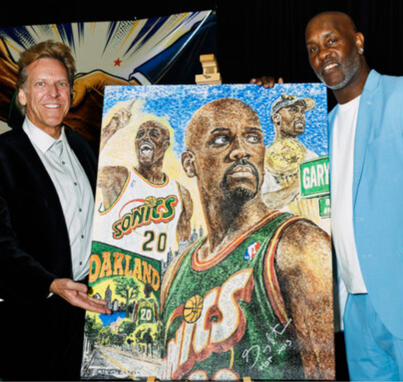 Two men stand beside a colorful painting of basketball players and team logos, holding the artwork, in an indoor setting.