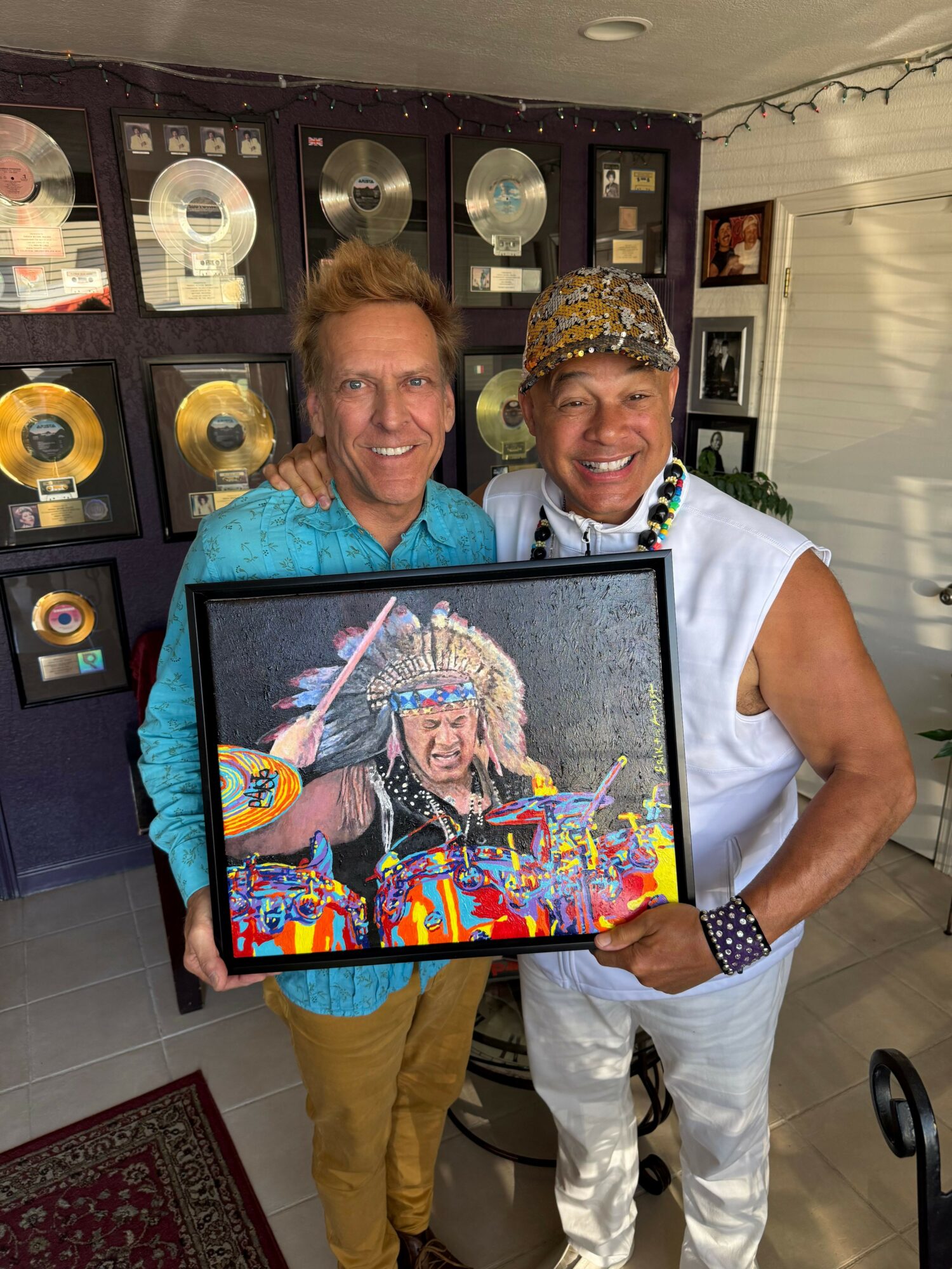 Two people smiling, one holding a framed colorful artwork, standing in front of a wall with gold records.