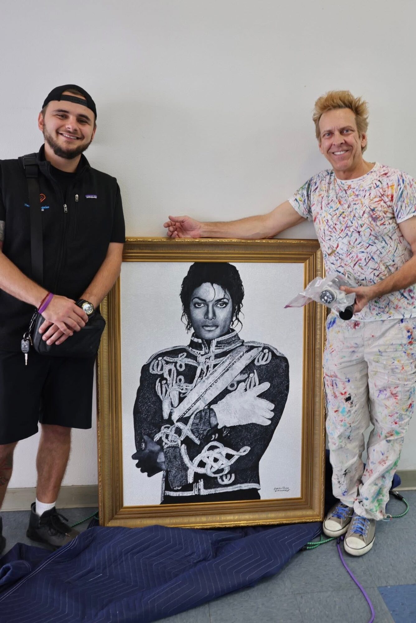 Two men stand beside a framed black-and-white portrait of a woman holding a guitar, in a room with a plain wall.
